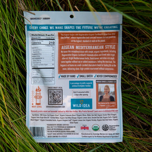 Back of a bag of Agavean Mediterranean Style Pickled Beets, photographed outdoors on green grass. The packaging is white with blue, orange, and red text blocks, showing nutritional facts, ingredients, and a small photo of a person.