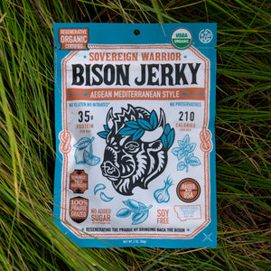 A package of Sovereign Warrior Bison Jerky, AegEAN Mediterranean Style, placed on a bed of green grass. The packaging is bright blue with white and orange text and graphics, featuring a stylized bison head illustration.
