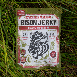 A package of Sovereign Warrior Bison Jerky, Bulgogi Korean BBQ flavor, placed flat on a bed of green grass. The packaging is olive green with a large white label featuring a black bison illustration and red text.