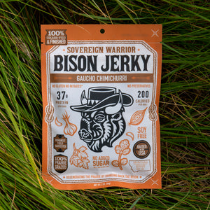A package of Sovereign Warrior Bison Jerky, Gaucho Chimichurri flavor, placed flat on a background of green grass. The packaging is orange and white with a central illustration of a bison wearing a cowboy hat.