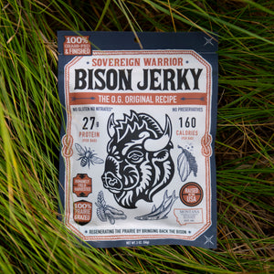 A package of Sovereign Warrior Bison Jerky is placed flat on a bed of green grass. The package is dark blue with a large white and orange label featuring a black illustration of a bison.