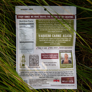 The back of a Wild Idea Vaquero Carne Asada beef jerky package is photographed lying on green grass outdoors. The package is rectangular and white, with large brown and green text blocks, a nutrition facts panel, a QR code, and a small photo of a smiling man.