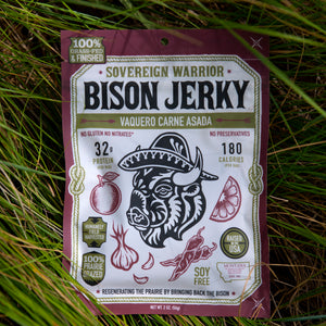 A package of Sovereign Warrior Bison Jerky, specifically Vaquero Carne Asada, lying flat on a bed of green grass. The packaging is a deep maroon with a large white central label featuring a black graphic of a bison wearing a sombrero, surrounded by red and green illustrations of ingredients like an apple, garlic, and chili peppers.