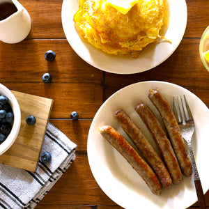 A breakfast table scene with two white plates on a wooden table: one plate holds a golden omelet with butter, and the other holds four browned sausage links with a fork. Blueberries, a small pitcher, and a napkin are also visible in a styled overhead composition.
